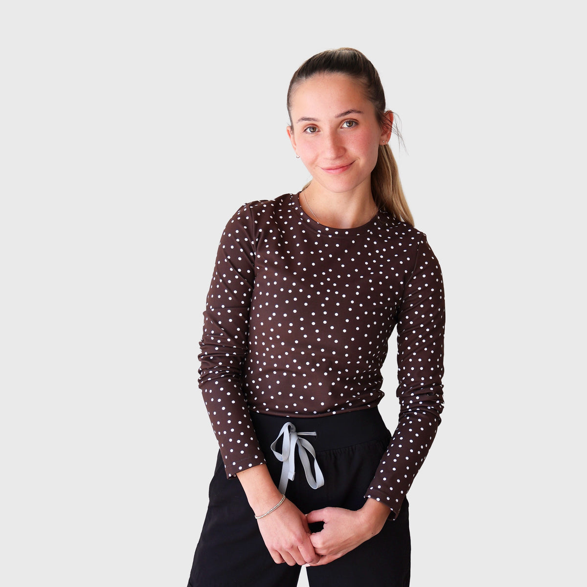 Chocolate PolkaDot CoreFit Underscrub, front view: long-sleeve crew-neck top in chocolate brown with small white polka dots, worn with black scrub pants