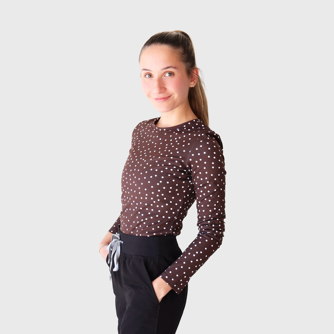 Chocolate PolkaDot CoreFit Underscrub, front view: long-sleeve crew-neck top in chocolate brown with small white polka dots, worn with black scrub pants