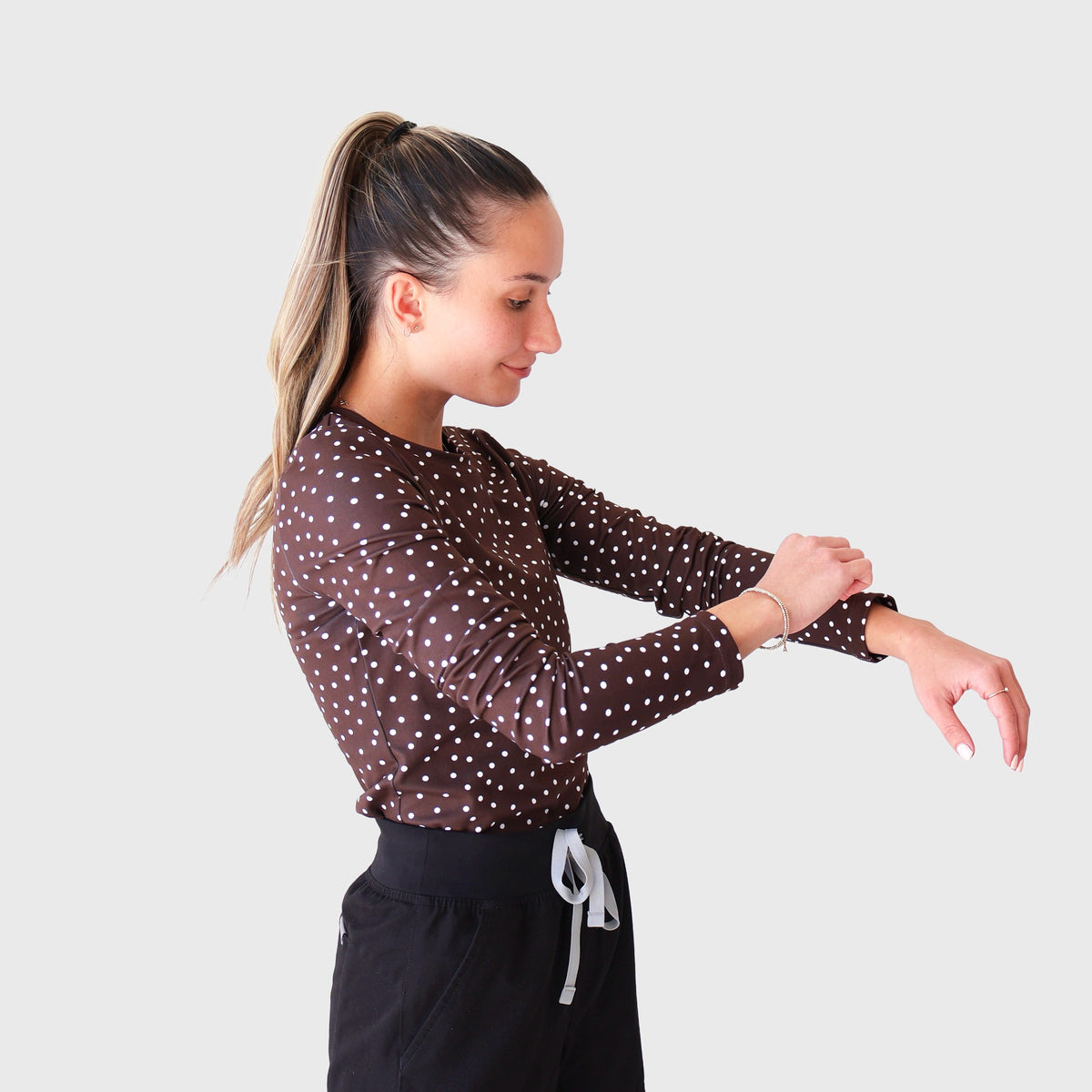 Chocolate PolkaDot CoreFit Underscrub, side view adjusting the cuff: long-sleeve chocolate brown top with small white polka dots