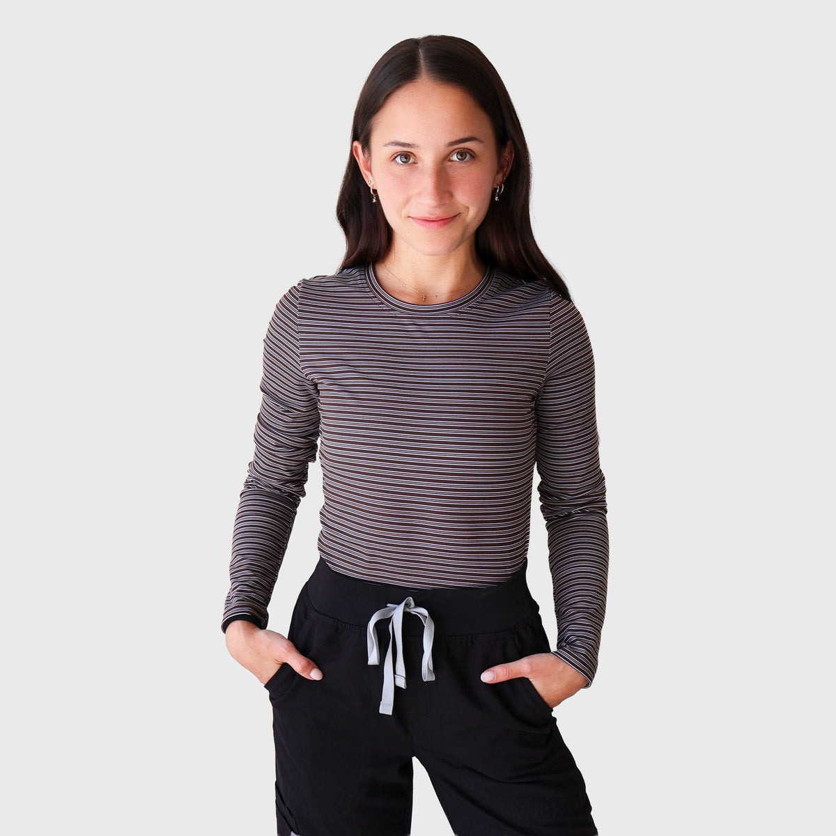 Fall Stripes CoreFit Underscrub, front view with hands in pockets: long-sleeve crew-neck top in fine taupe and black stripes, worn with black scrub pants